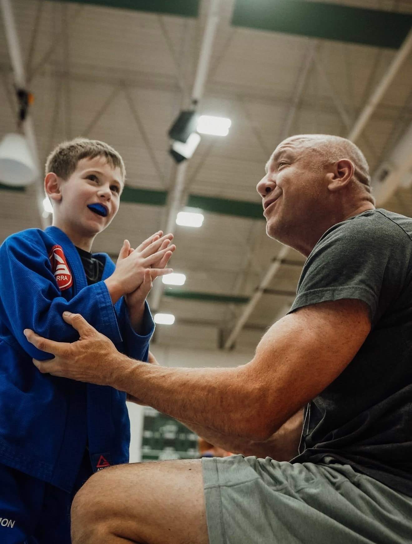 A boy in a blue Gracie Barra gi and mouthguard high-fives his coach