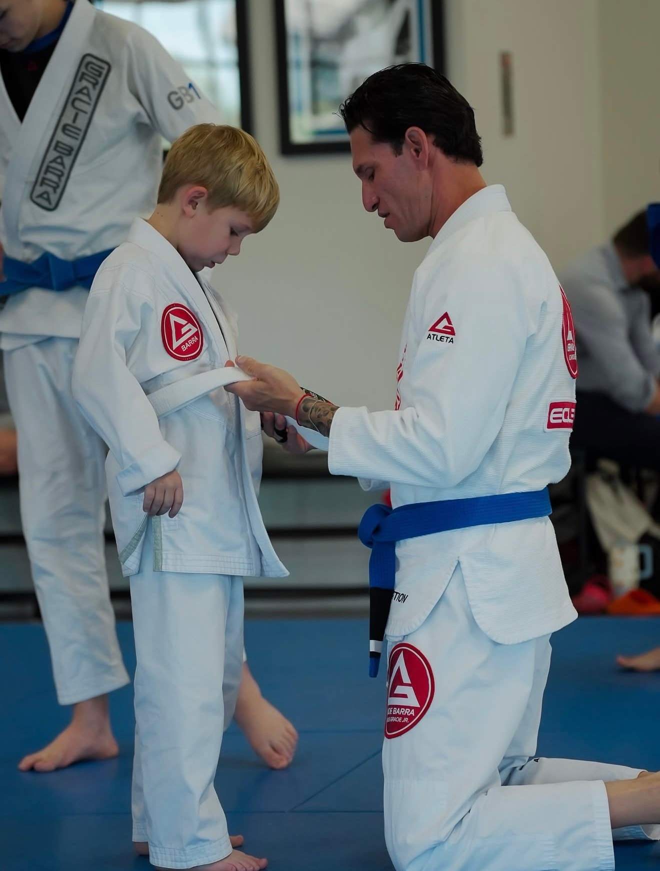 A coach kneels to tie the white belt on a young student’s gi at Gracie Barra Ocotillo