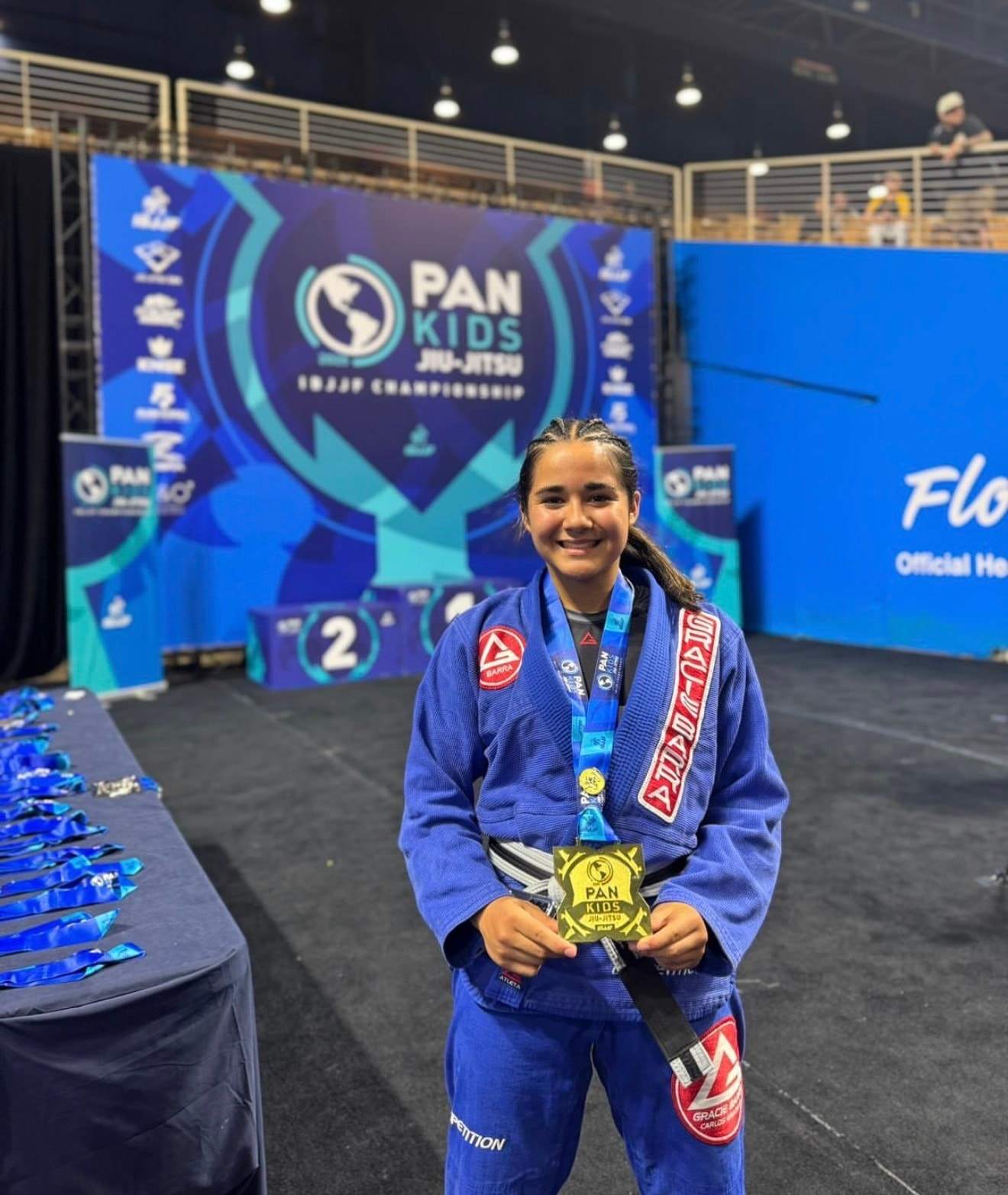 A smiling young competitor in a blue Gracie Barra gi holds a gold medal at the IBJJF Pan Kids Championship