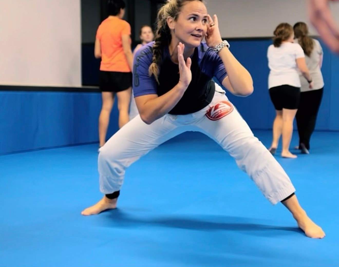 A woman in a rash guard and gi pants practices a defensive stance during a women’s class
